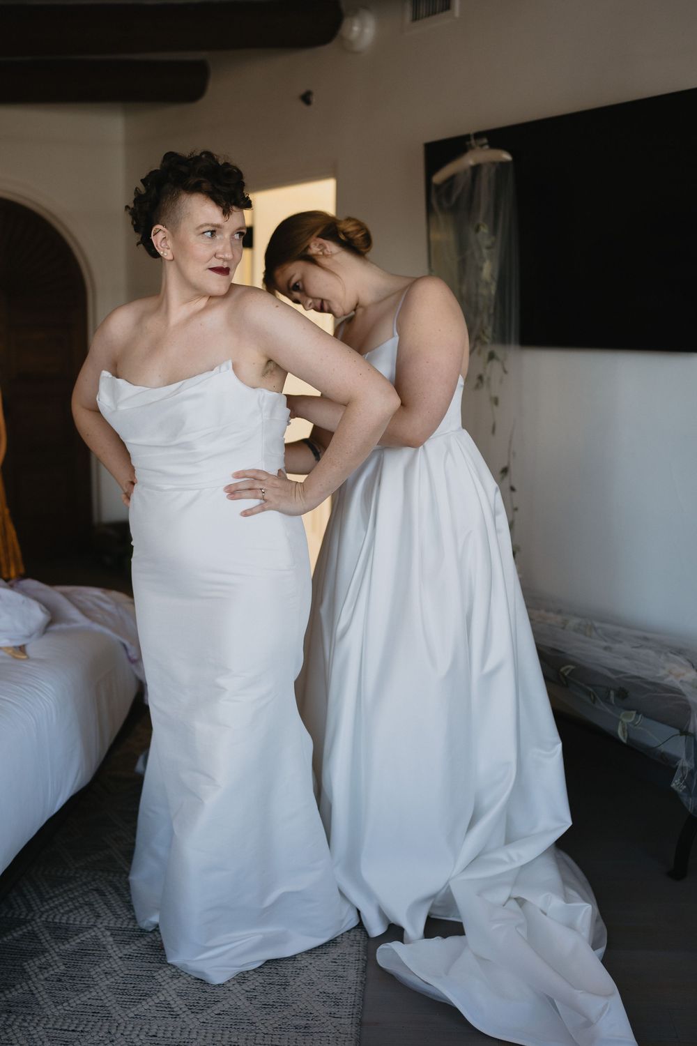 Two brides in white wedding gowns help each other get dressed in a bright hotel room before their ceremony.
