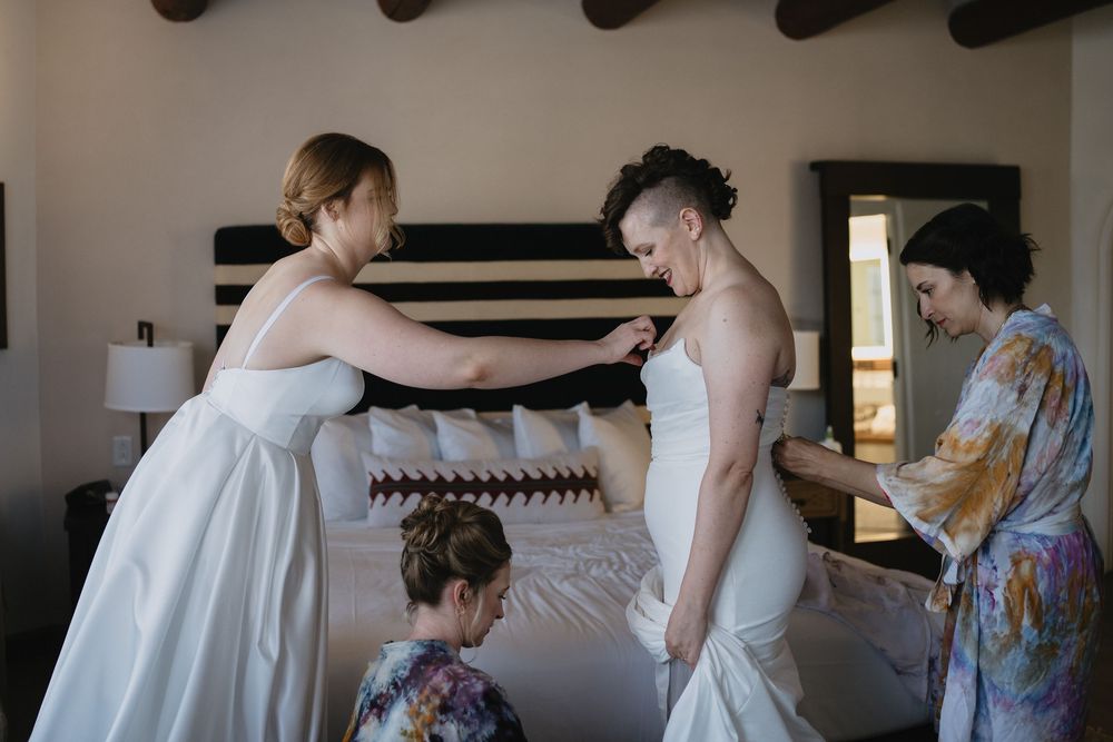 Bridesmaids helping bride get dressed in elegant bedroom, adjusting white wedding gown details before ceremony.