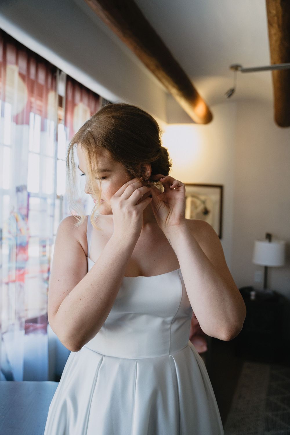 Bride in white dress putting on earrings by window with soft natural light on her wedding day.