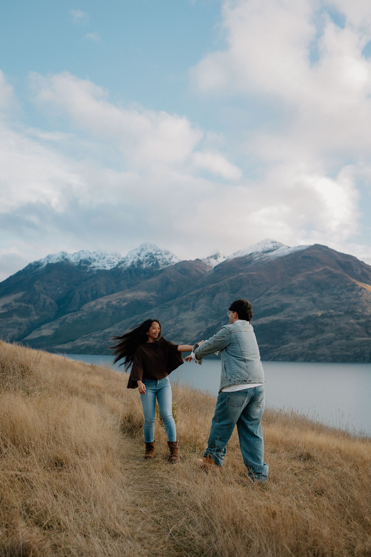 Woman and young girl walk hand-in-hand on gravel path with snow-capped mountains backdrop in winter.