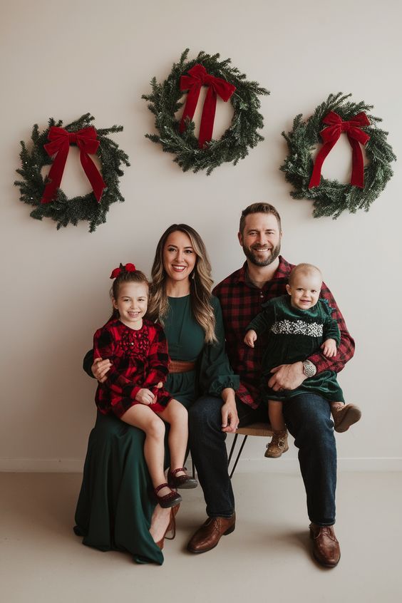 Family of four in red and green Christmas outfits posing with holiday wreaths on white wall backdrop.