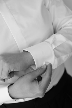 Close-up black and white photo of a groom fastening his cufflinks on a white dress shirt.
