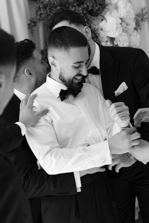 Black and white photo of groom laughing joyfully while groomsmen embrace him before wedding.