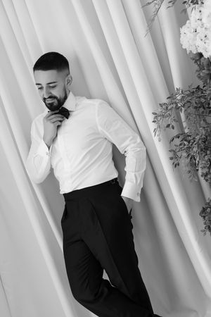 Elegant groom in white shirt and black trousers adjusting his bow tie near draped curtains.