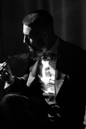 Dramatic black and white portrait of a bearded groom in a tuxedo smoking a cigar.