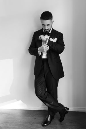 Dapper bearded groom in black tuxedo adjusting his cufflinks while leaning against a white wall.