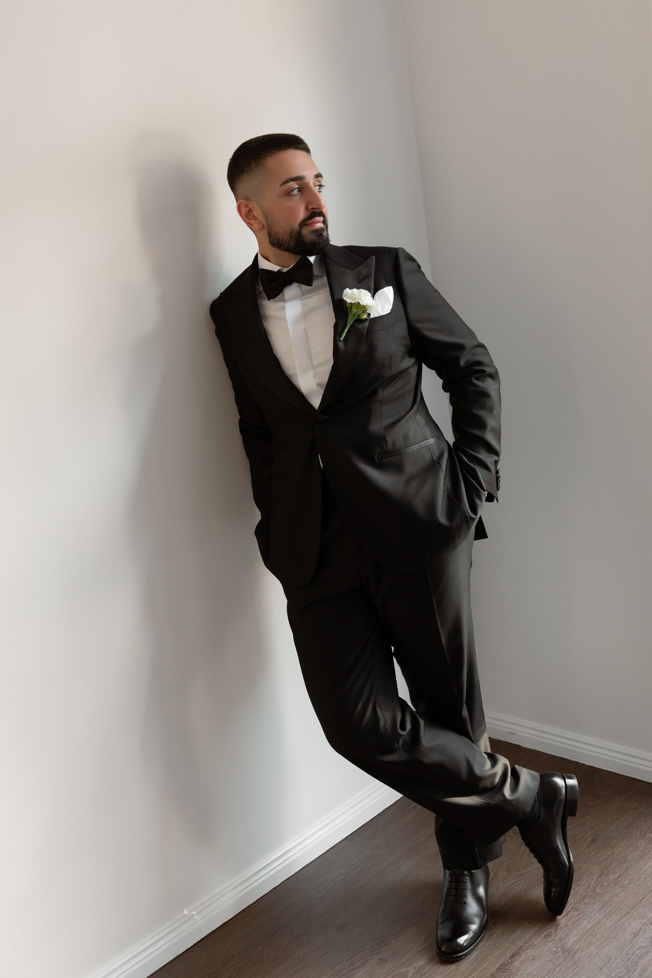 Stylish bearded groom in black tuxedo with white boutonniere leaning against a white wall.