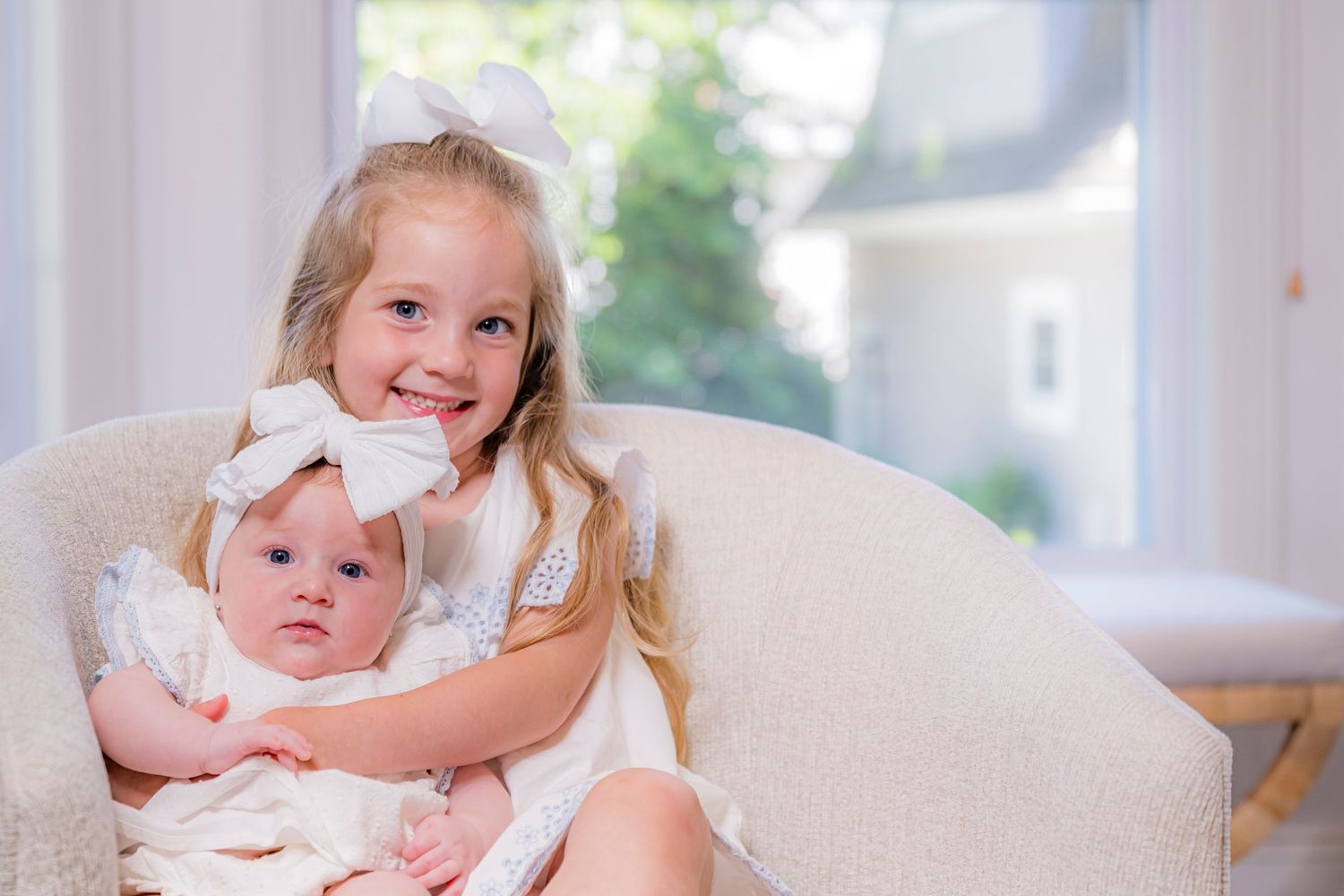 Young girl with white bow holds baby sister in cream chair by bright window, both wearing white dresses.