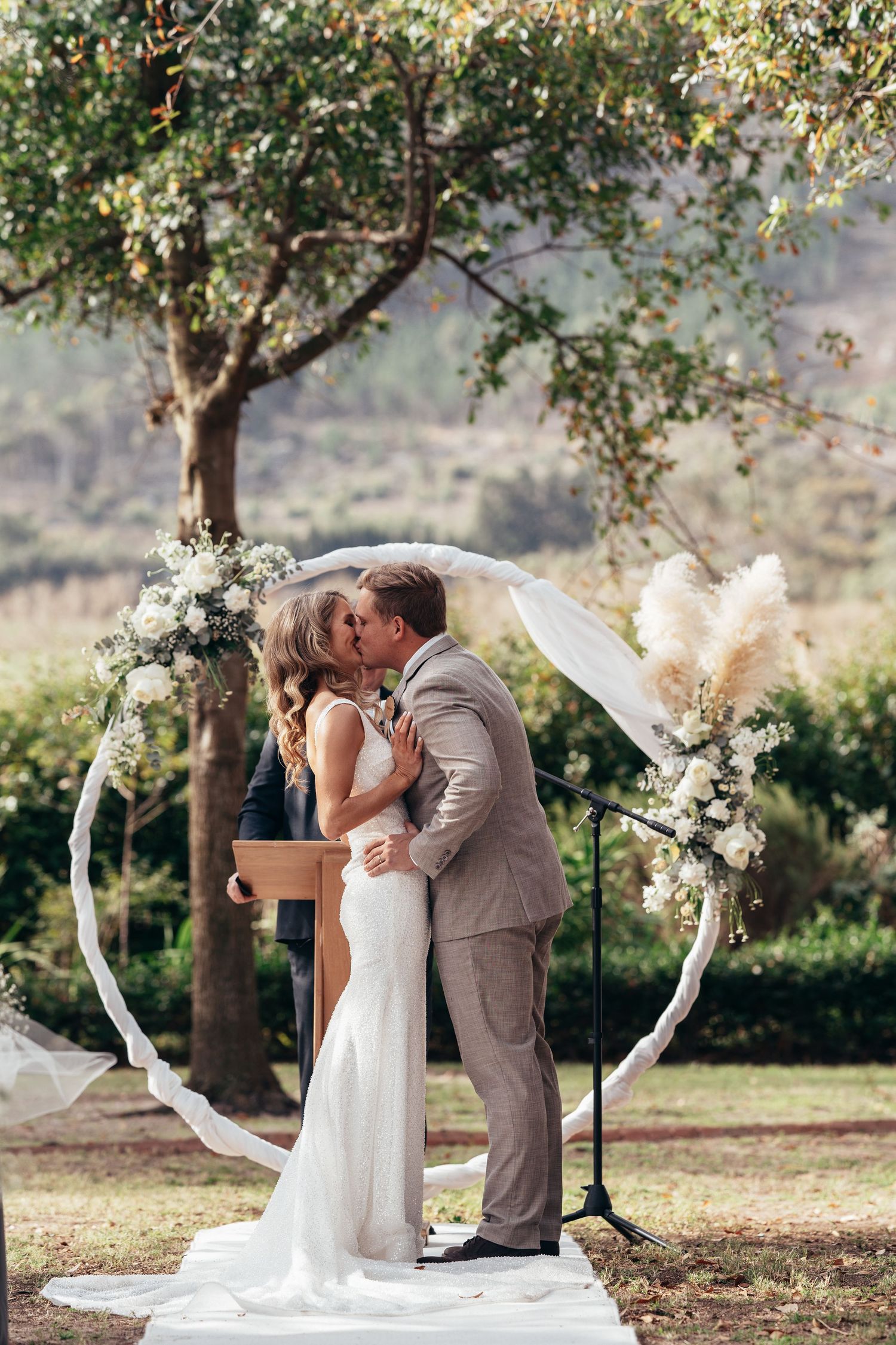 Bride and groom sharing first kiss under floral circular arch at outdoor wedding ceremony.