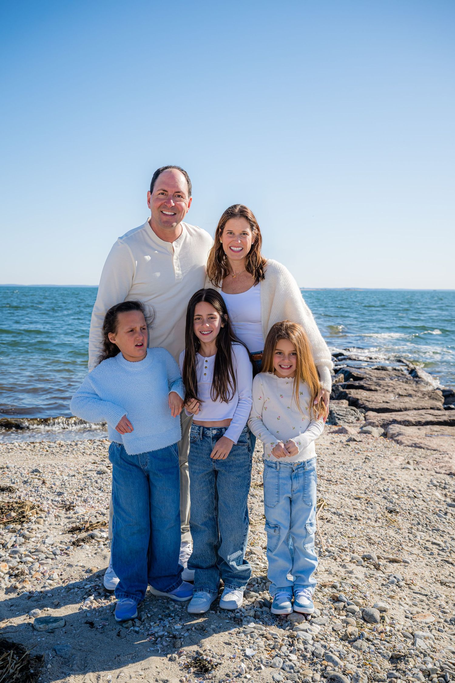 Happy family of five posing together on a rocky beach, wearing white sweaters and jeans on a sunny day.