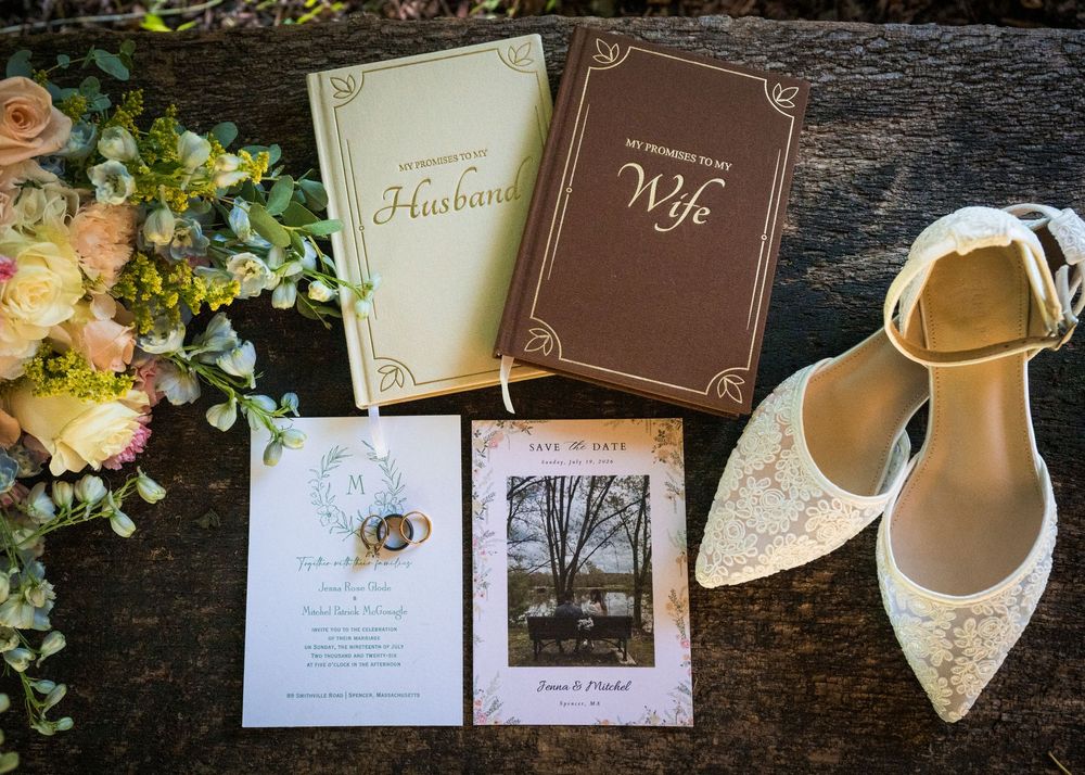 Wedding flat lay featuring bridal heels, floral bouquet, save the date cards, and husband and wife journals on rustic wood.