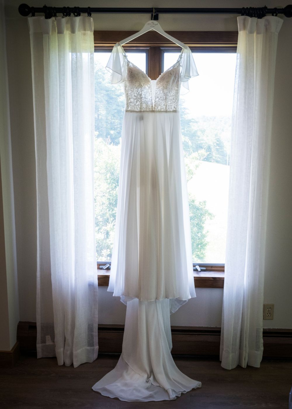 Elegant white wedding dress with lace bodice hanging in a sunlit window with sheer curtains.