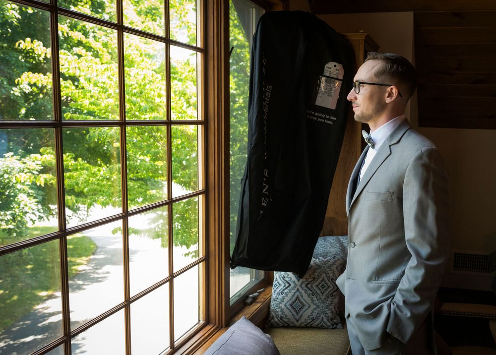 Man in gray suit and glasses gazes out large window at lush green trees, with garment bag hanging nearby.