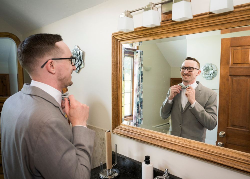 Groom in gray suit adjusting bow tie while looking in a wooden-framed bathroom mirror before wedding ceremony.