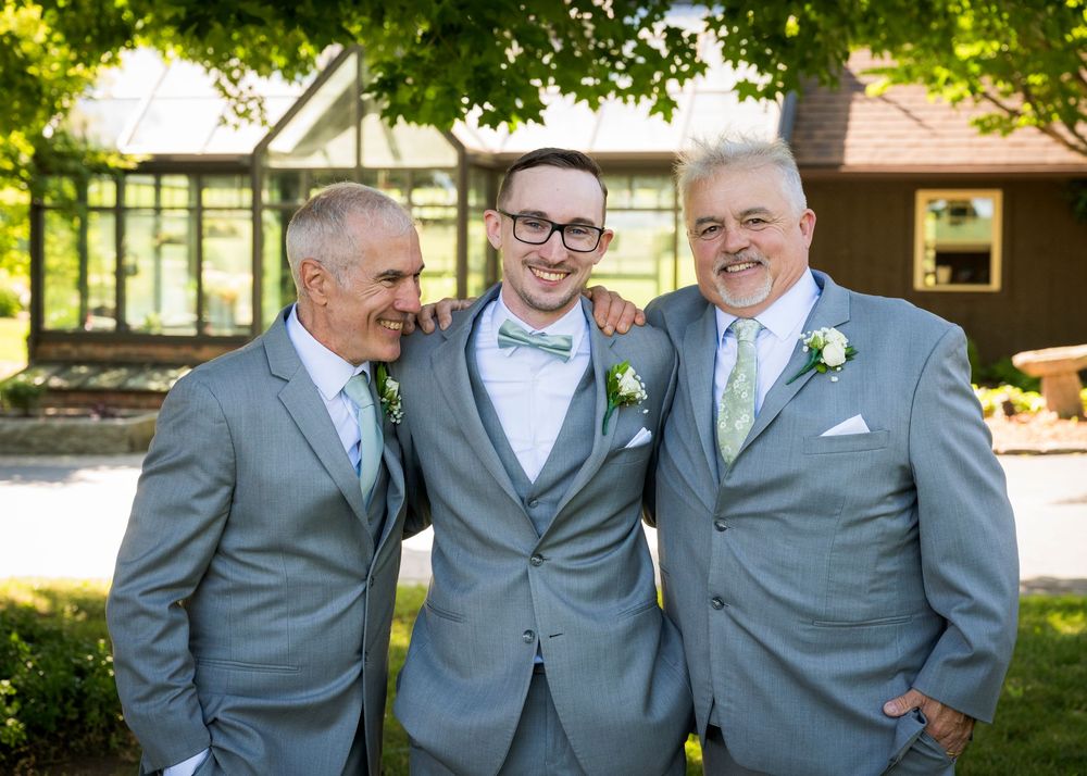 Three men in matching gray suits with green ties smile together outdoors at a wedding celebration.