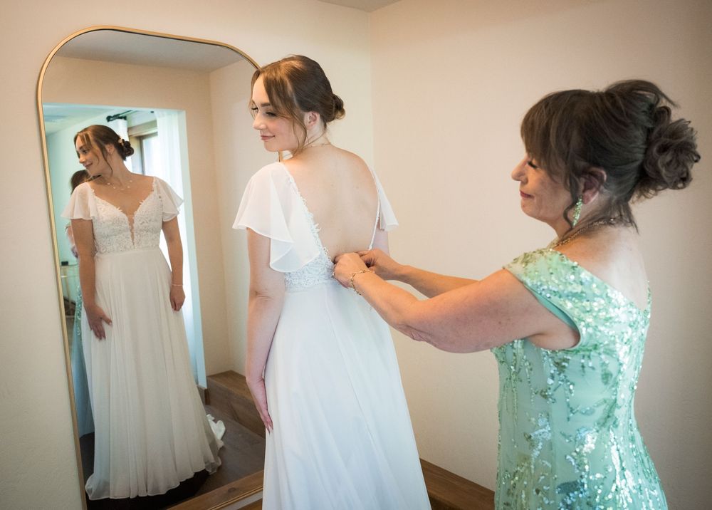 Mother helping bride button her elegant white wedding gown while reflected in a full-length mirror.