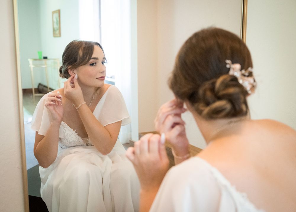 Bride in white dress adjusting earrings while looking in mirror on wedding day.