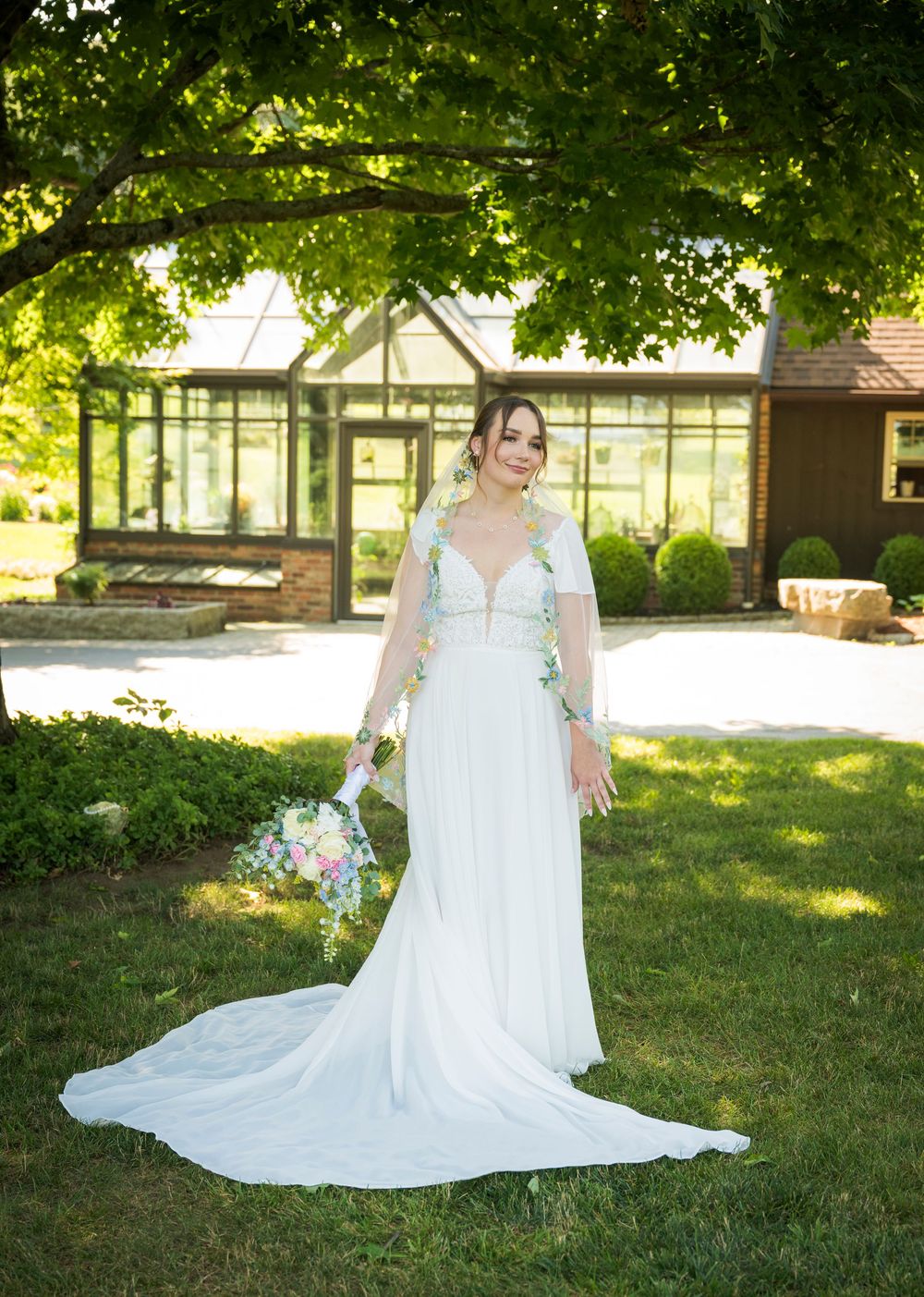 Bride in a flowing white wedding gown with floral sleeves holding a bouquet outdoors near a greenhouse.