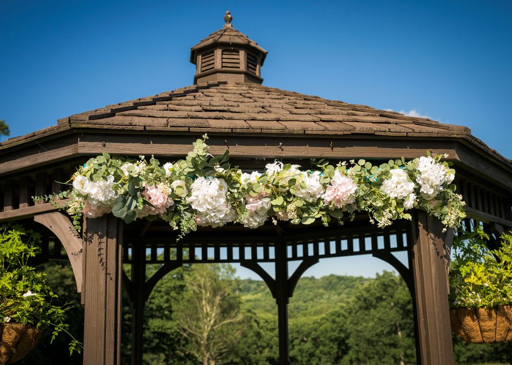 Brown wooden gazebo adorned with lush floral garland of pink roses and greenery against a clear blue sky.