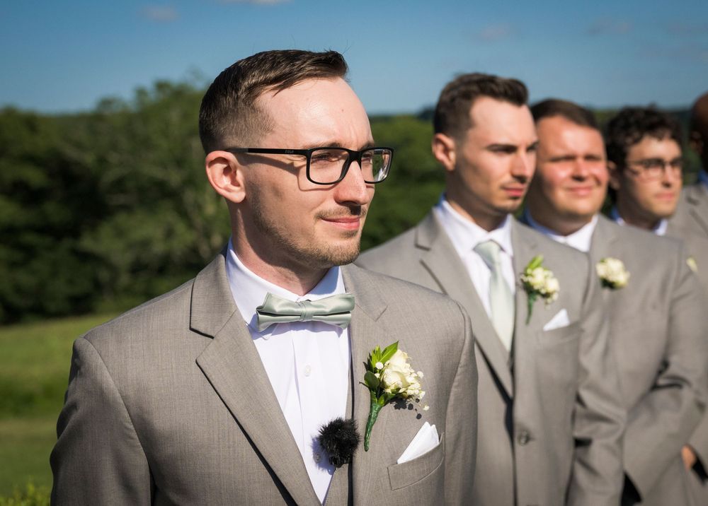 Groom in gray suit with bow tie and boutonniere stands with groomsmen outdoors at a wedding ceremony.