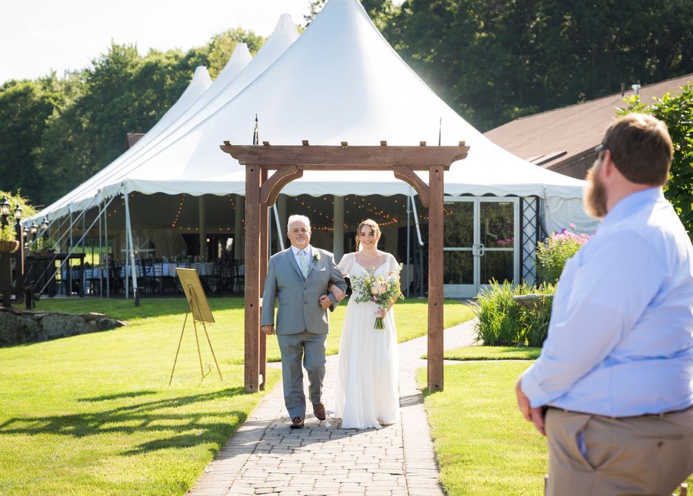 Bride and groom walking down outdoor wedding aisle under wooden arbor with white tent and lush green lawn.