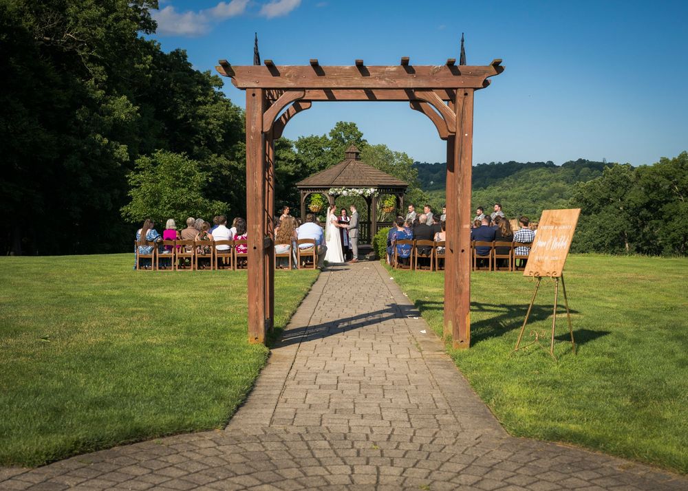 Outdoor wedding ceremony under a rustic wooden pergola arch on a brick path surrounded by lush green hills.