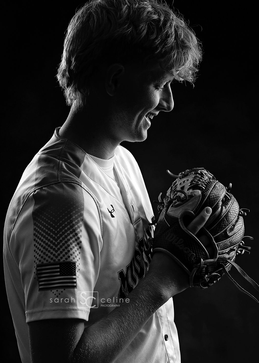 Black and white senior portrait of a smiling baseball player in uniform holding a glove, dramatic studio lighting.