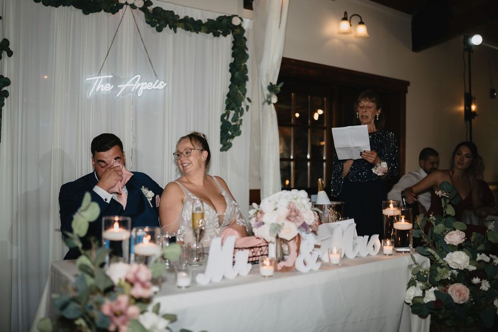 Bride and groom laughing at wedding reception head table with floral arrangements and neon sign backdrop.