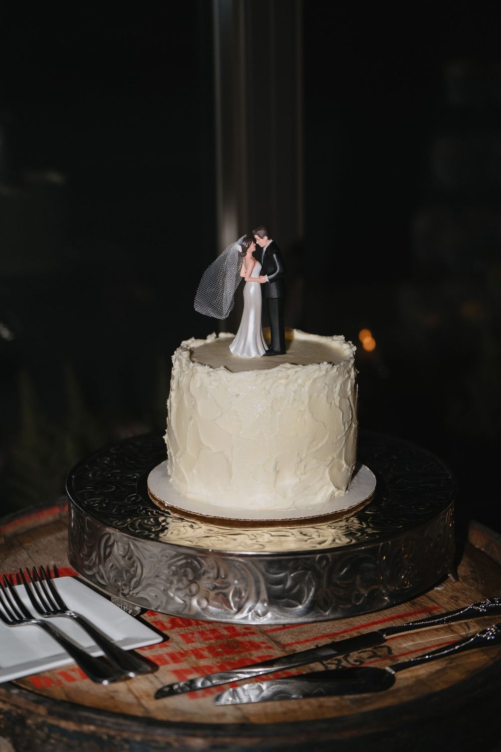 Single-tier white wedding cake with bride and groom topper on silver stand, surrounded by serving utensils.