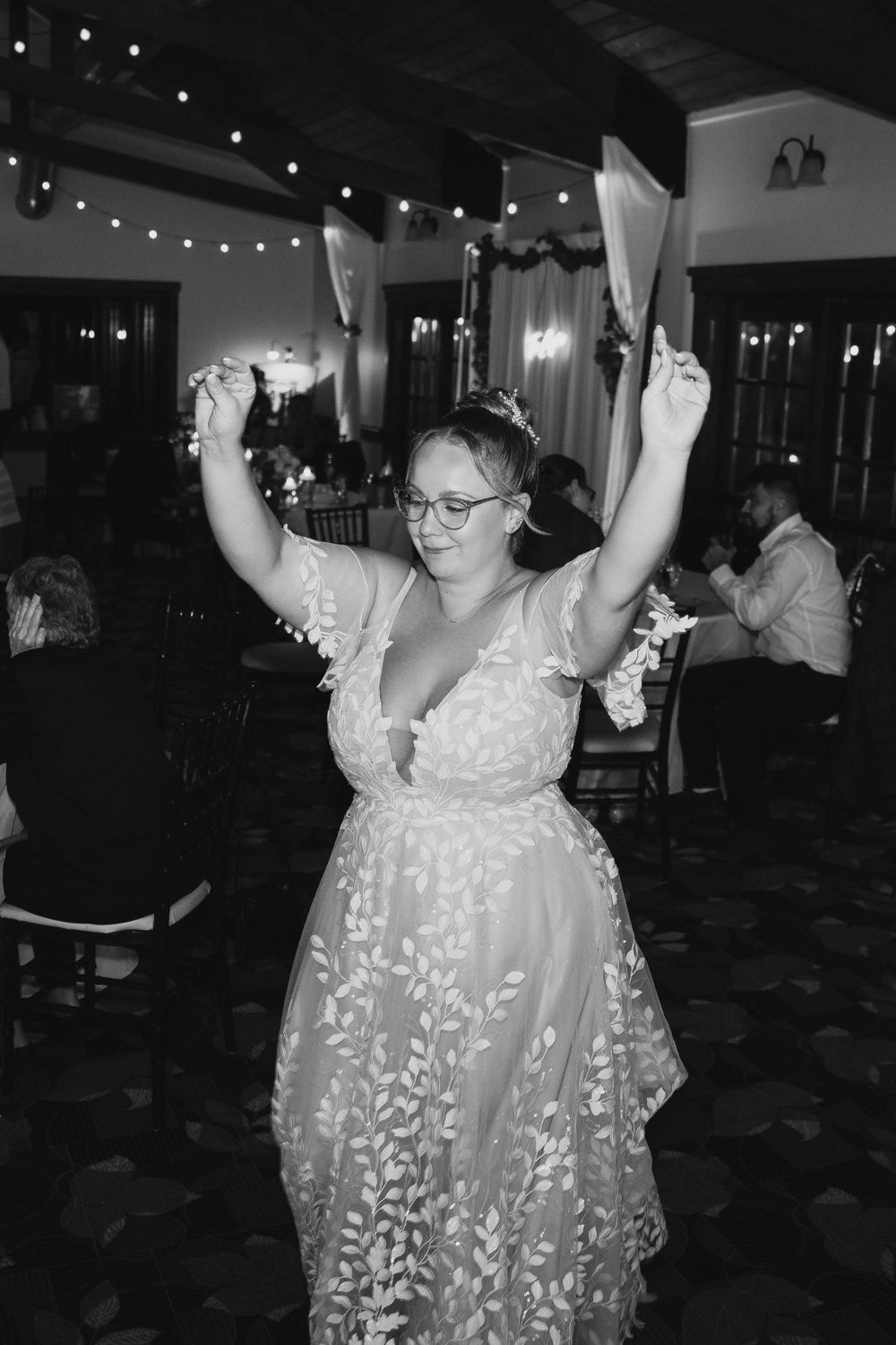 Woman in floral gown dancing joyfully at indoor wedding reception with string lights in black and white photo.