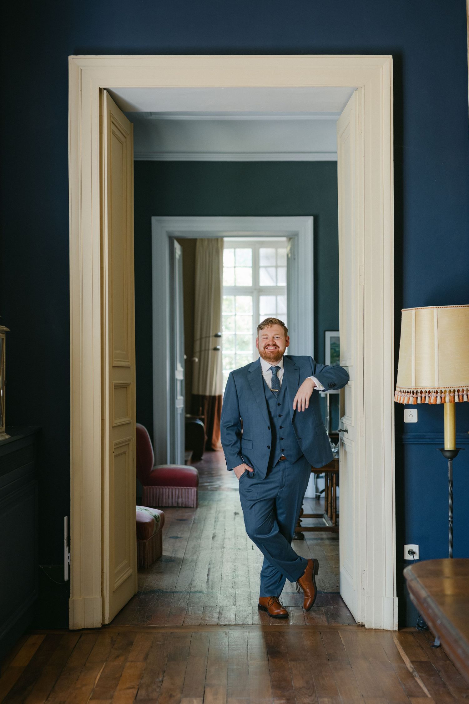 Man in blue suit leaning against doorframe in elegant dark navy room with hardwood floors.