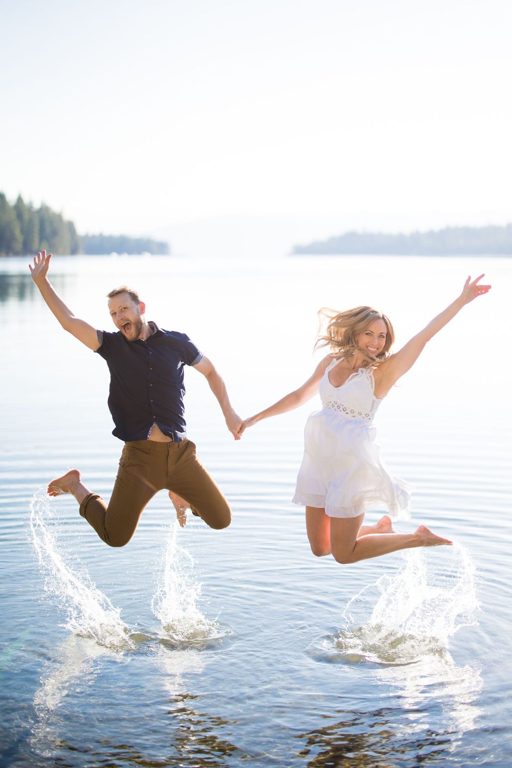 Joyful couple jumping together over a lake, holding hands, splashing water, wearing casual summer outfits on a sunny day.
