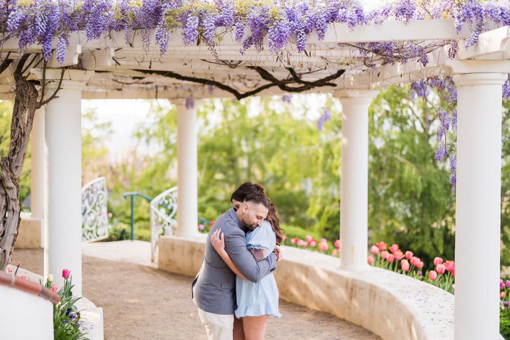 Couple embracing under wisteria-covered pergola with white columns and colorful tulips in a romantic garden setting.