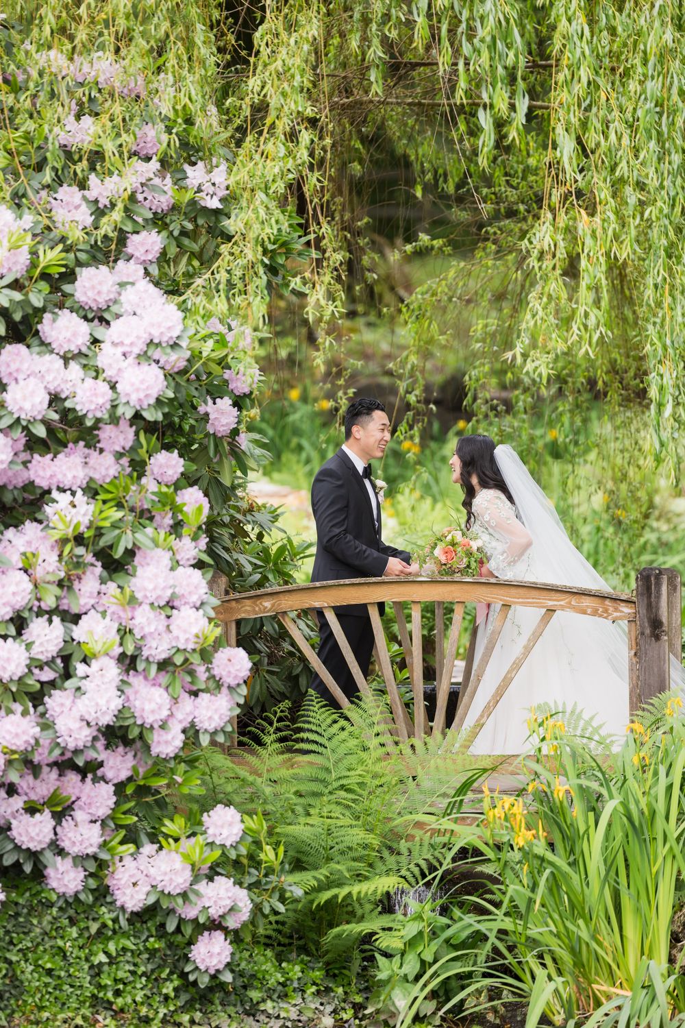 Bride and groom stand on a wooden bridge surrounded by lush greenery and blooming pink rhododendrons in a garden.