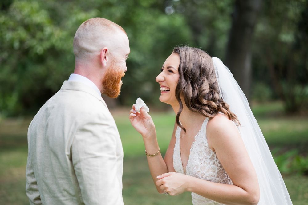 Bride laughing joyfully while feeding cake to groom with red beard outdoors at garden wedding ceremony.