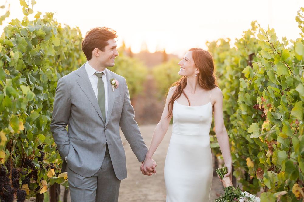 Bride and groom hold hands walking through a sunlit vineyard, bride in white gown, groom in gray suit.
