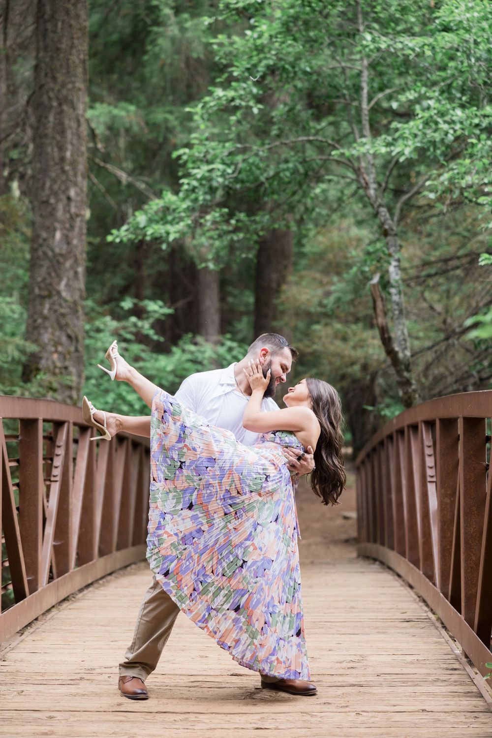 Couple sharing a romantic dip kiss on a wooden bridge surrounded by lush green forest trees.
