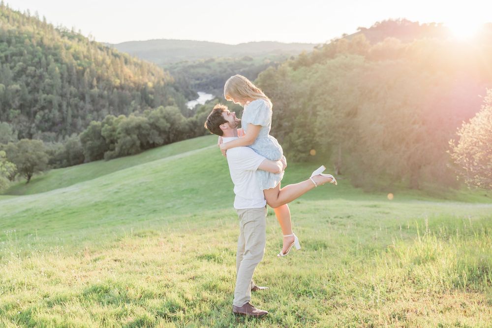 Couple sharing a romantic kiss at sunset in a green meadow, man lifting woman in his arms with hills in background.