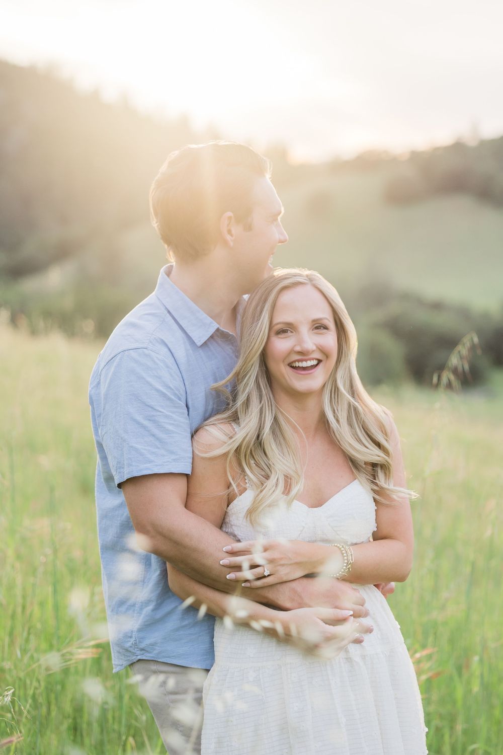 Couple embracing in sunlit green meadow, woman in white dress laughing joyfully, golden hour engagement photo.