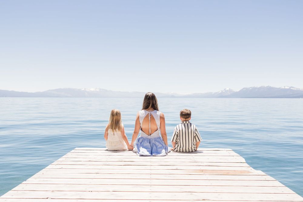 Woman and two children sitting on a wooden dock overlooking a calm lake with mountains in the background.