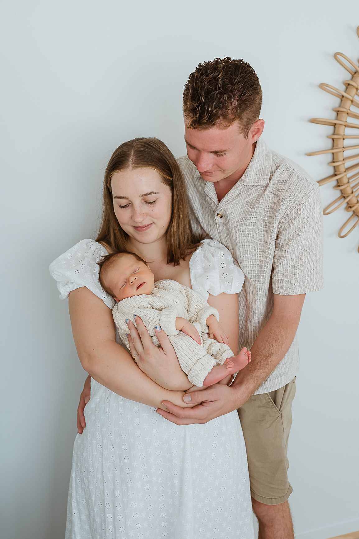 New parents lovingly cradle their sleeping newborn baby, dressed in cream outfits, against a soft light blue wall.