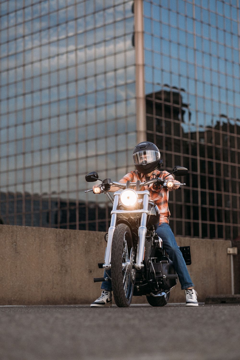 A motorcyclist in a helmet and plaid shirt sits on a classic motorcycle with headlight on, parked in front of a glass building.