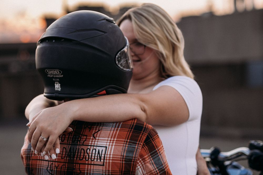 A woman in a white shirt hugs a person wearing a Harley-Davidson flannel and motorcycle helmet at sunset.