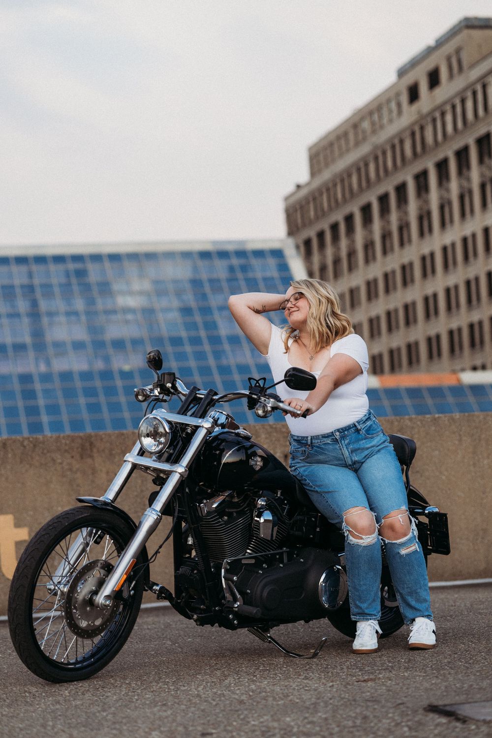 A smiling blonde woman in a white top and ripped jeans leans against a black Harley-Davidson motorcycle on a rooftop.