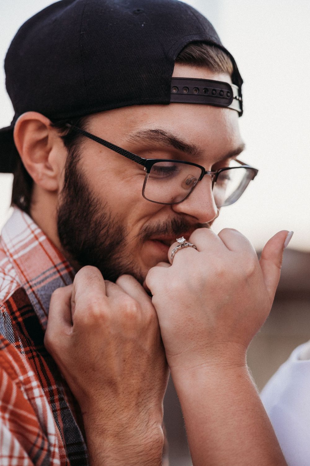 Bearded man in glasses and snapback cap kissing a woman's hand with an engagement ring.