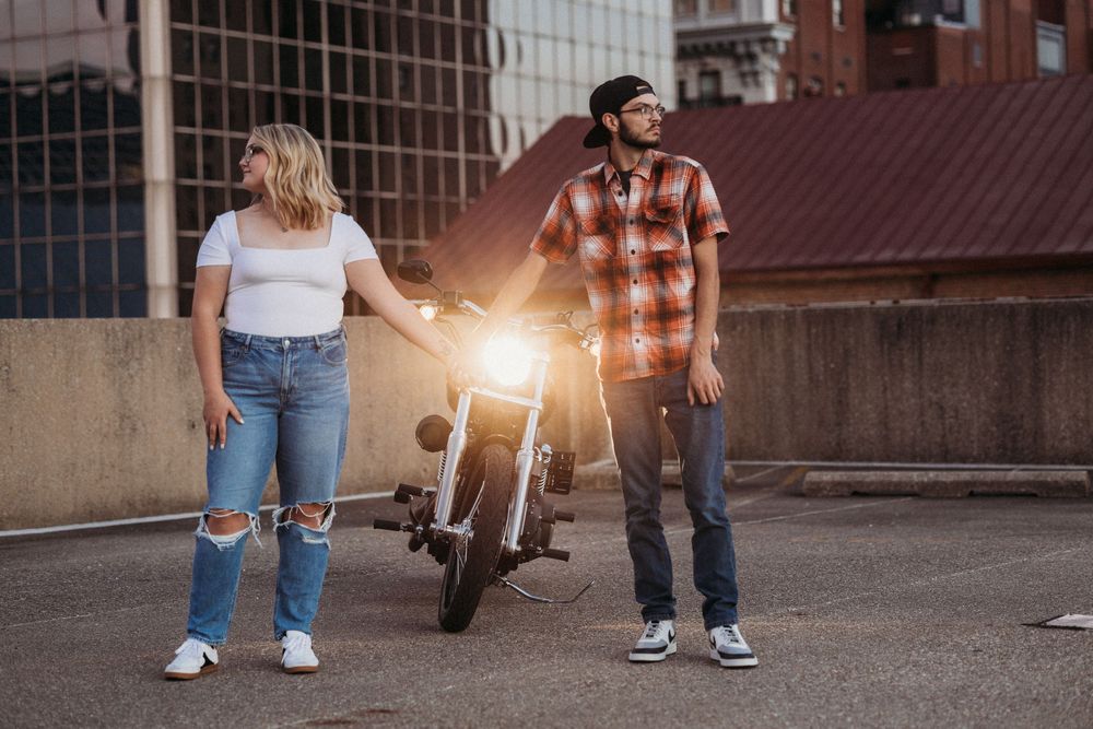 A man in a plaid shirt and helmet stands with a woman in jeans beside a motorcycle on an urban street.