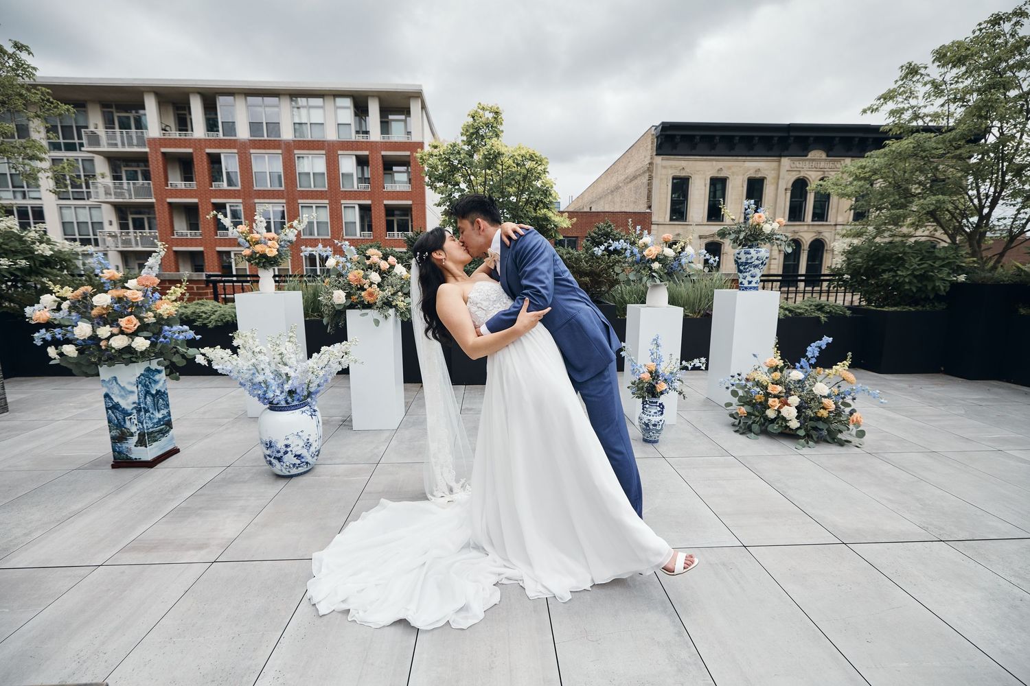 Bride and groom share a romantic dip kiss on a rooftop terrace surrounded by elegant floral arrangements on white pedestals.