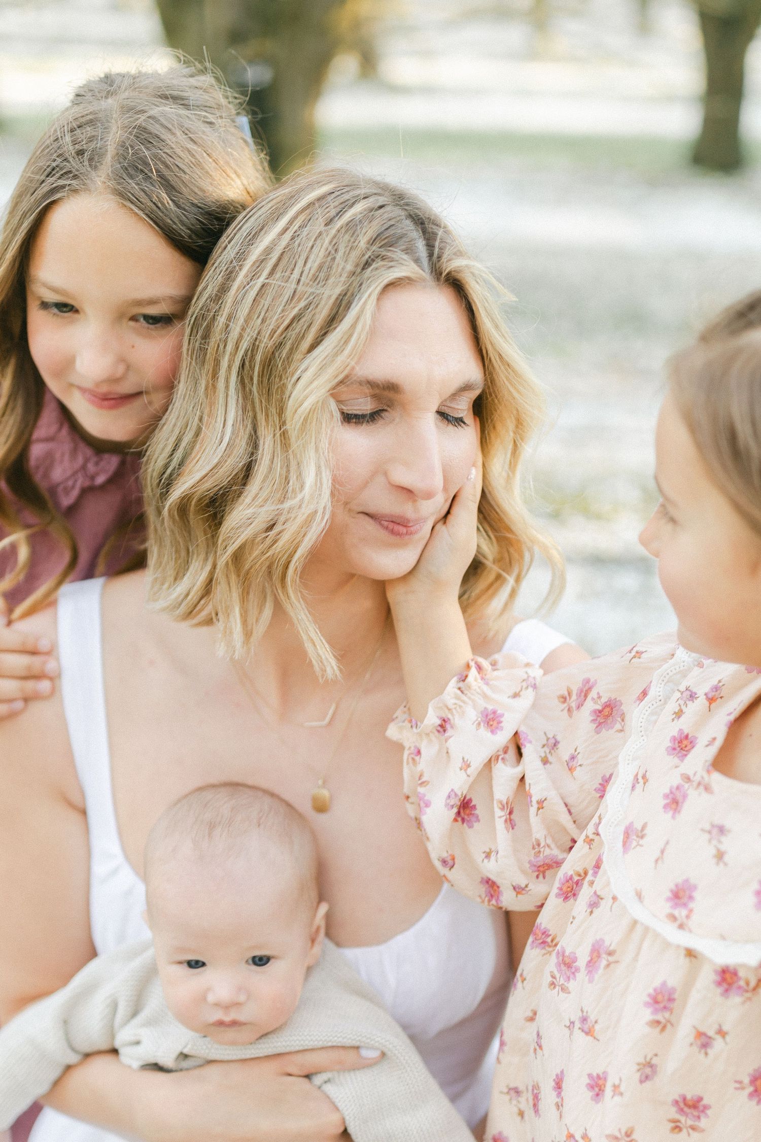 A mother holds her newborn baby while two young daughters lovingly surround her outdoors in soft natural light.