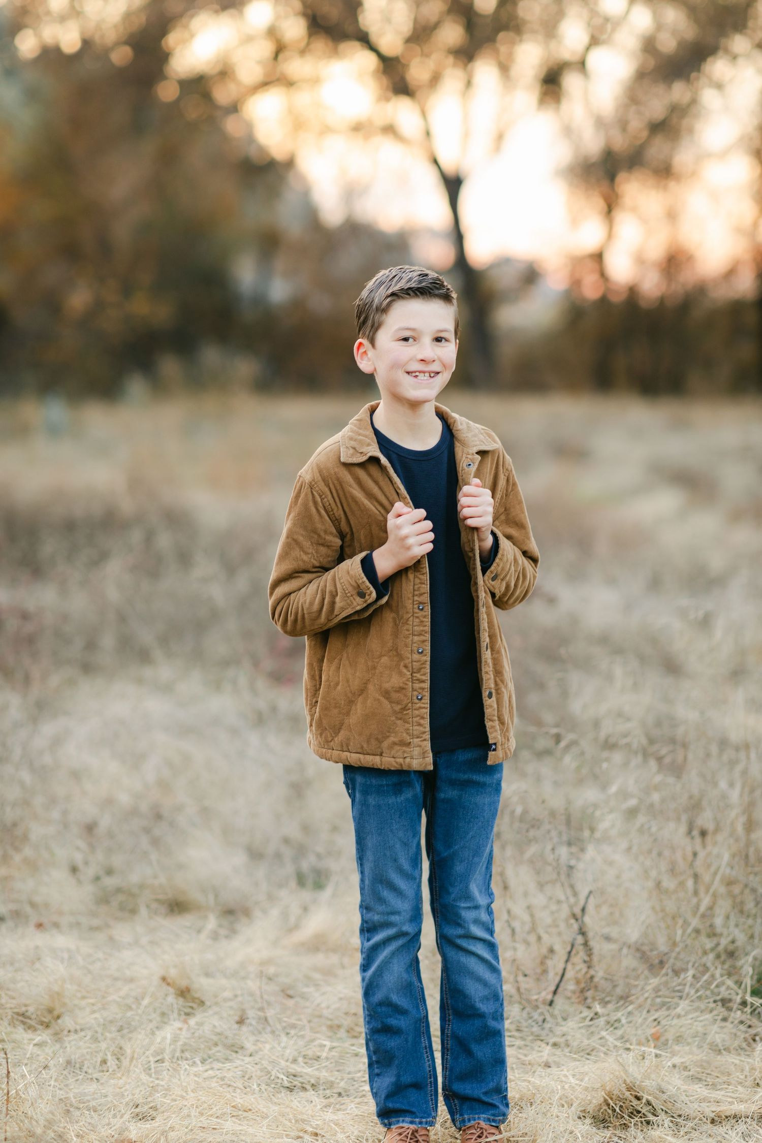 Smiling boy in brown corduroy jacket and jeans standing in a golden autumn field at sunset.