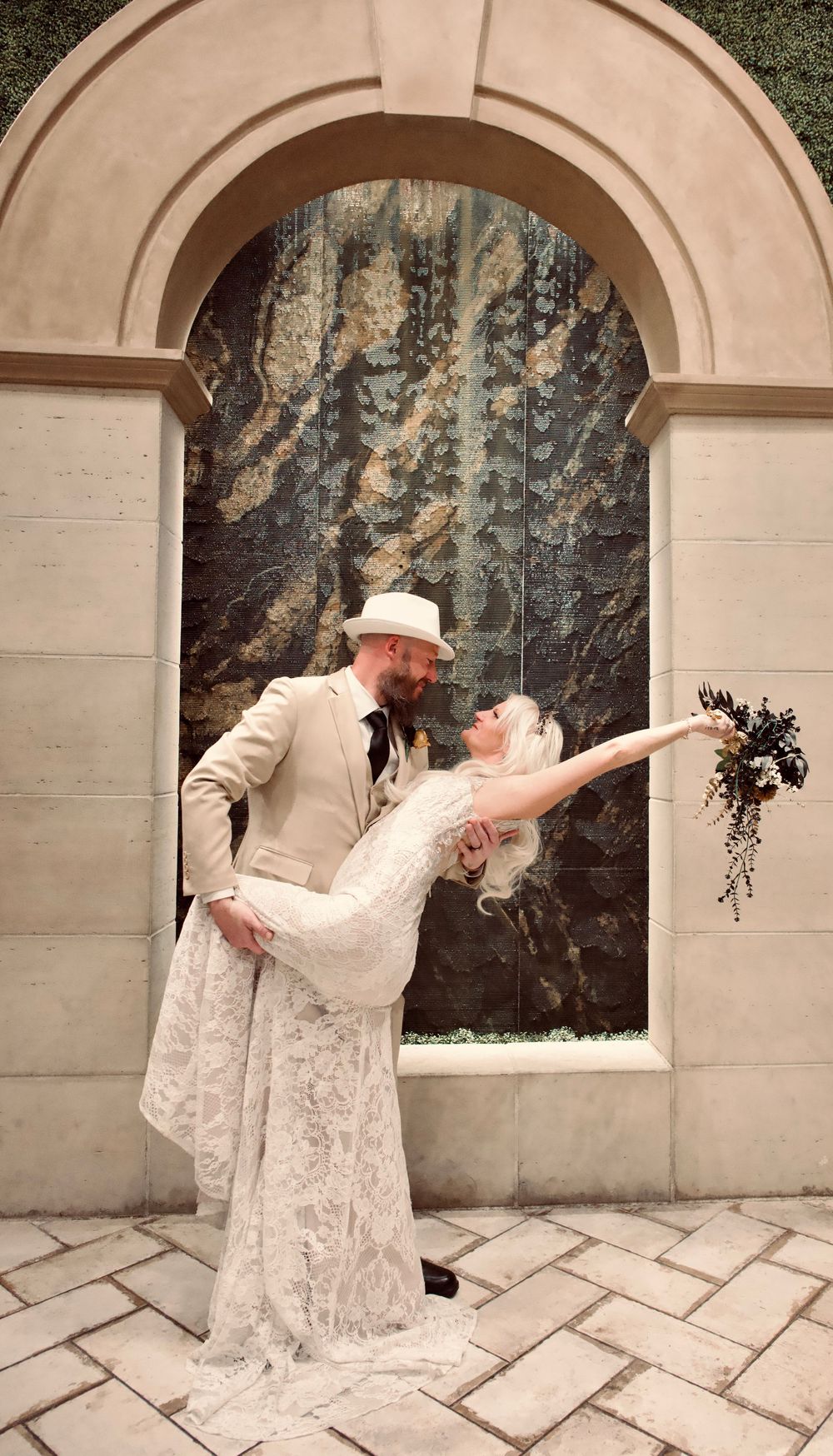 Groom dipping bride in a romantic pose before an arched stone wall, bride holding a cascading dark floral bouquet.